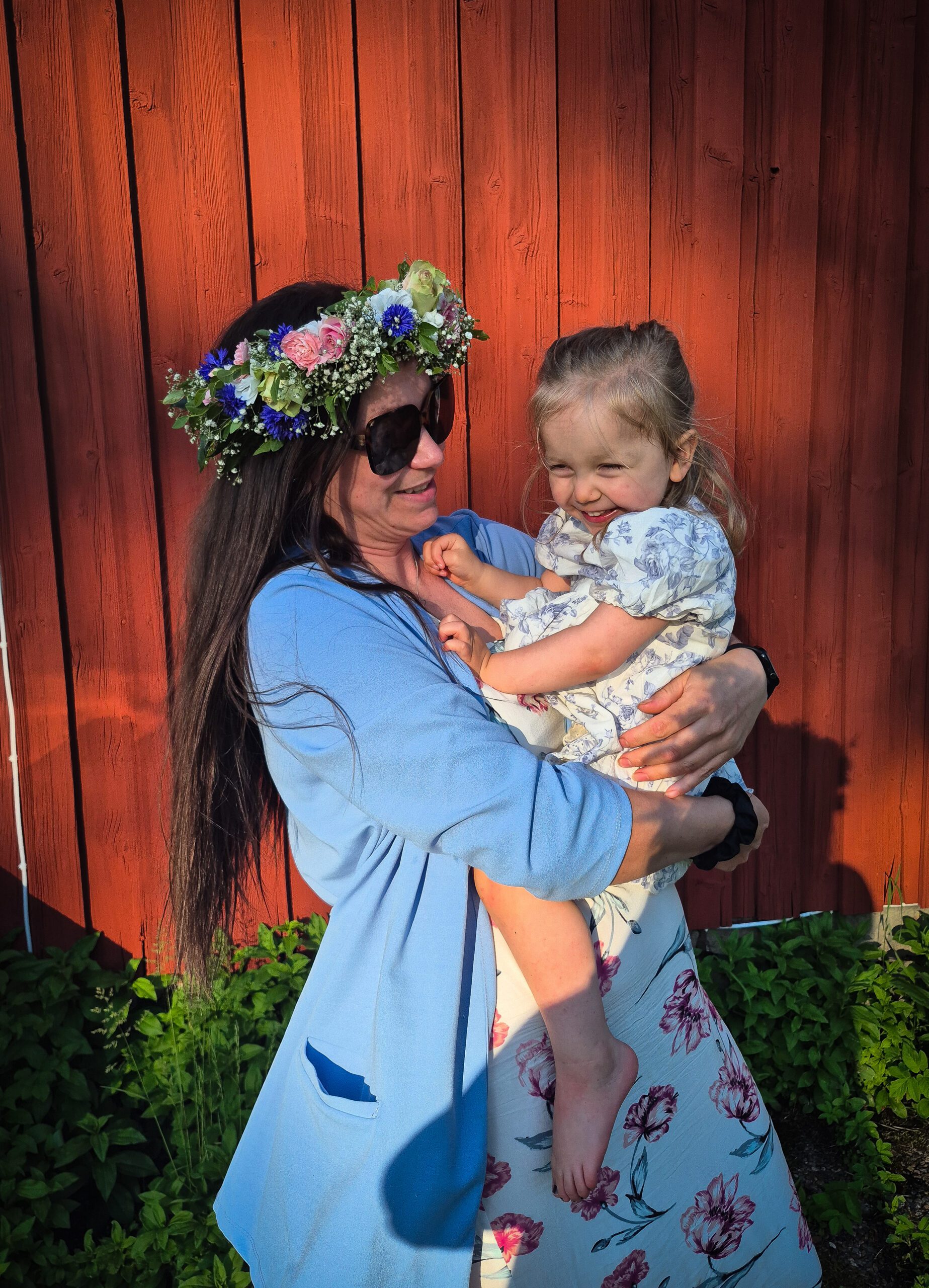 Jennifer håller Hilma i famnen framför en röd husvägg en sommardag.